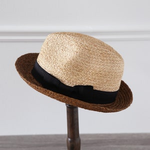 May include: A brown and beige straw fedora hat with a black ribbon. The hat is sitting on a wooden stand.