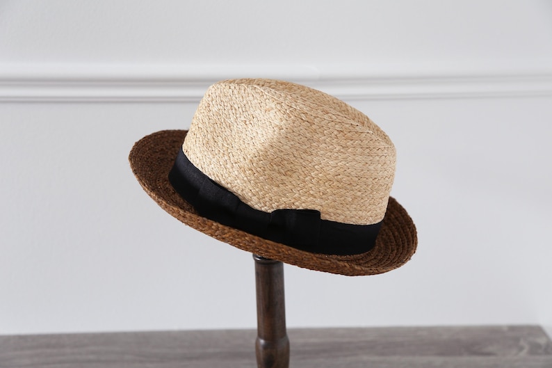 May include: A brown and beige straw fedora hat with a black ribbon. The hat is sitting on a wooden stand.