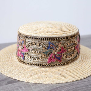 May include: A straw hat with a wide brim and a flat crown. The hat features a decorative band with colorful floral embroidery, sequins, and gold-colored trim. The hat is a light beige color.