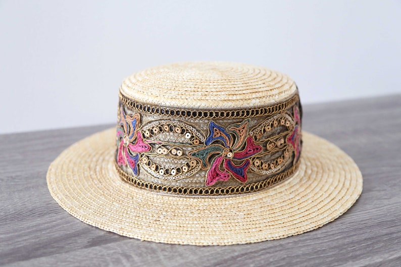 May include: A straw hat with a wide brim and a flat crown. The hat features a decorative band with colorful floral embroidery, sequins, and gold-colored trim. The hat is a light beige color.