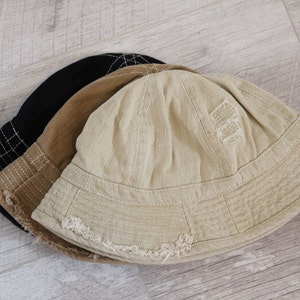 May include: Three bucket hats stacked on top of each other. The top hat is beige with a distressed look and a small embroidered design. The middle hat is brown and the bottom hat is black.