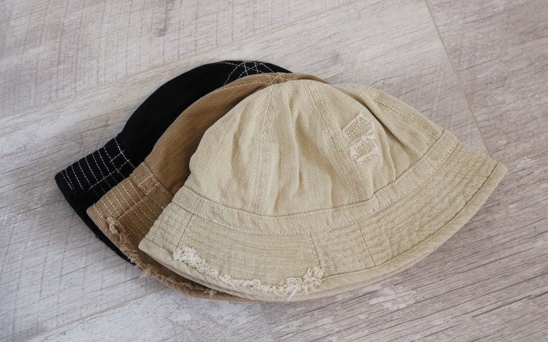 May include: Three bucket hats stacked on top of each other. The top hat is beige with a distressed look and a small embroidered design. The middle hat is brown and the bottom hat is black.