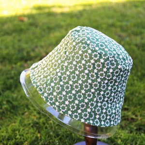 May include: A green bucket hat with a white floral pattern. The hat is on a clear display stand.
