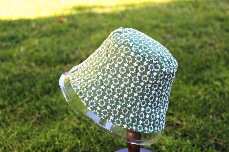 May include: A green bucket hat with a white floral pattern. The hat is on a clear display stand.