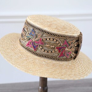 May include: A straw hat with a wide brim and a flat crown. The hat features a decorative band with floral embroidery in pink, blue, and green, accented with gold sequins and trim. The hat is displayed on a wooden stand.