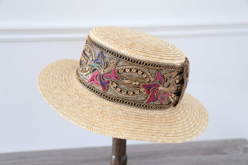 May include: A straw hat with a wide brim and a flat crown. The hat features a decorative band with floral embroidery in pink, blue, and green, accented with gold sequins and trim. The hat is displayed on a wooden stand.