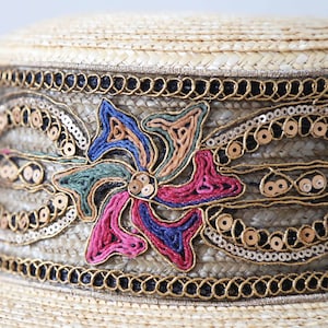 May include: A straw hat with a decorative band. The band features a floral design with pink, blue, and green embroidery, gold sequins, and gold trim. The hat has a natural straw color.