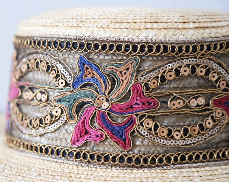May include: A straw hat with a decorative band. The band features a floral design with pink, blue, and green embroidery, gold sequins, and gold trim. The hat has a natural straw color.