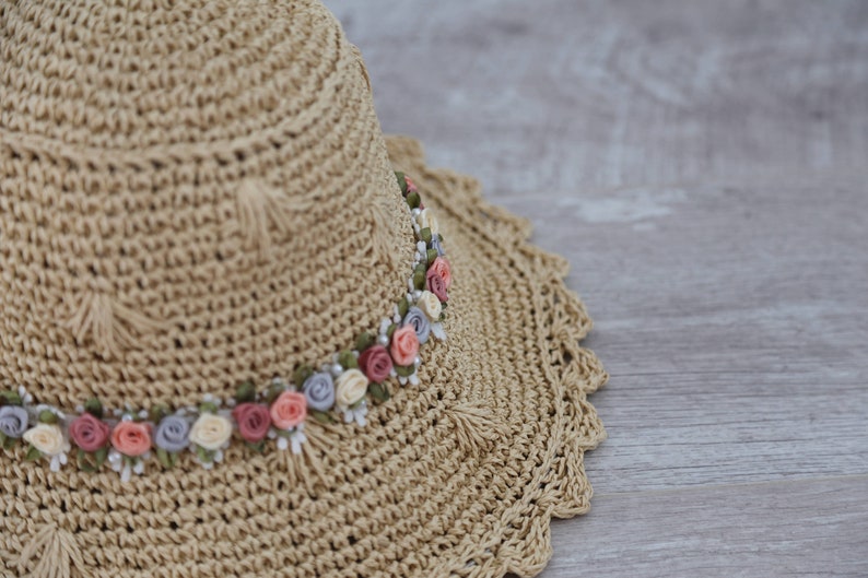 Puede incluir: Un sombrero de paja con un borde de crochet y una banda de flores de tela en tonos de rosa, blanco y azul.