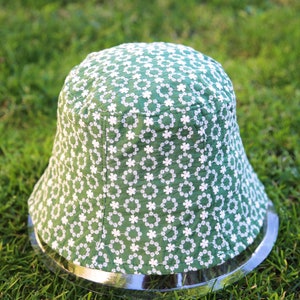 May include: A green bucket hat with a white floral pattern. The hat has a wide brim and a silver metal band around the bottom.