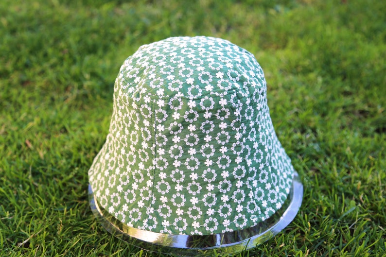 May include: A green bucket hat with a white floral pattern. The hat has a wide brim and a silver metal band around the bottom.
