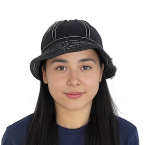 Puede incluir: Una mujer con el pelo largo y negro lleva un sombrero negro de cubo con costuras blancas. Lleva una camisa azul oscuro.