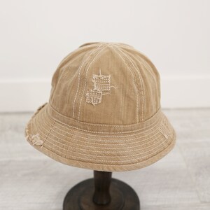 May include: A beige bucket hat with a distressed look and white stitching. The hat has a small patch of fabric missing on the crown.