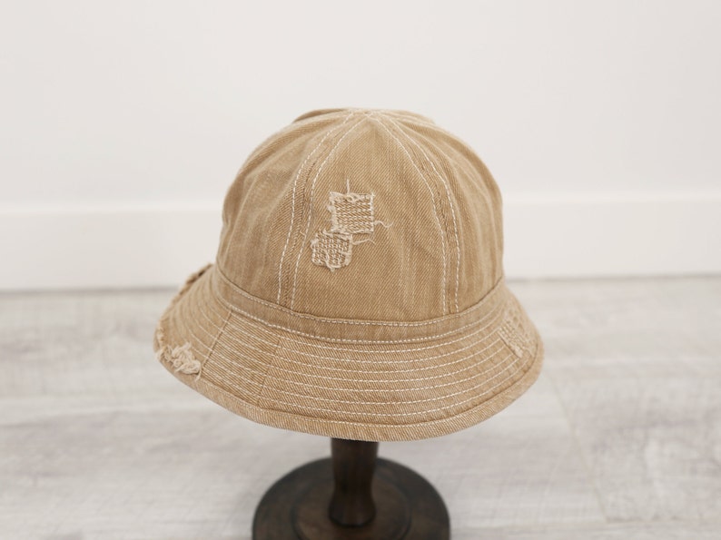 May include: A beige bucket hat with a distressed look and white stitching. The hat has a small patch of fabric missing on the crown.