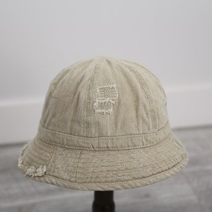 May include: A beige bucket hat with a distressed look. The hat has a wide brim and a slightly rounded crown. The hat is made of a soft, lightweight fabric.