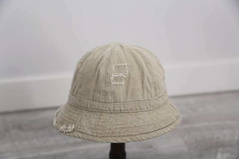 May include: A beige bucket hat with a distressed look. The hat has a wide brim and a slightly rounded crown. The hat is made of a soft, lightweight fabric.