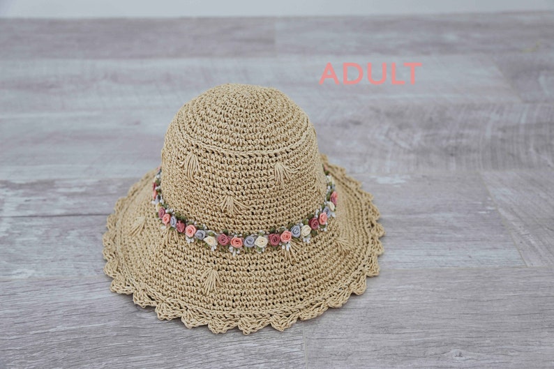 Puede incluir: Un sombrero de sol de crochet beige con un borde festoneado y una banda de flores rosas, blancas y moradas.