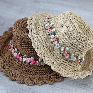 Puede incluir: Dos sombreros de paja tejidos a crochet con un adorno floral. El sombrero de arriba es beige con un borde marr&oacute;n y el sombrero de abajo es marr&oacute;n con un borde marr&oacute;n. Ambos sombreros tienen una banda de flores de tela rosa, blanca y azul claro.