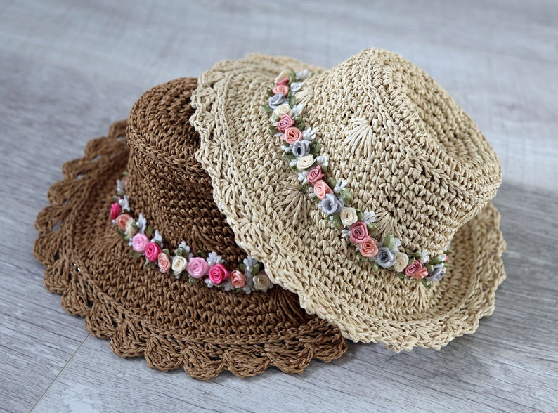 Puede incluir: Dos sombreros de paja tejidos a crochet con un adorno floral. El sombrero de arriba es beige con un borde marr&oacute;n y el sombrero de abajo es marr&oacute;n con un borde marr&oacute;n. Ambos sombreros tienen una banda de flores de tela rosa, blanca y azul claro.