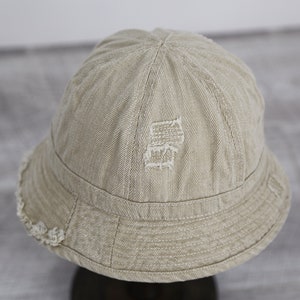 May include: A beige bucket hat with a distressed, embroidered design on the crown. The hat has a frayed edge around the brim.