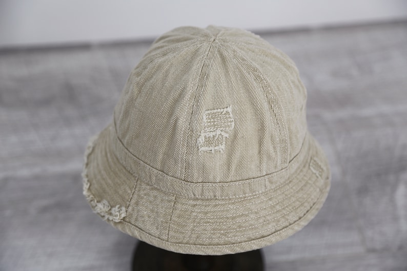 May include: A beige bucket hat with a distressed, embroidered design on the crown. The hat has a frayed edge around the brim.