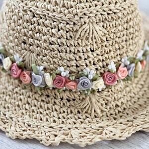 Puede incluir: Un sombrero de paja de ala ancha con una banda de flores de tela rosa, blanca y gris.