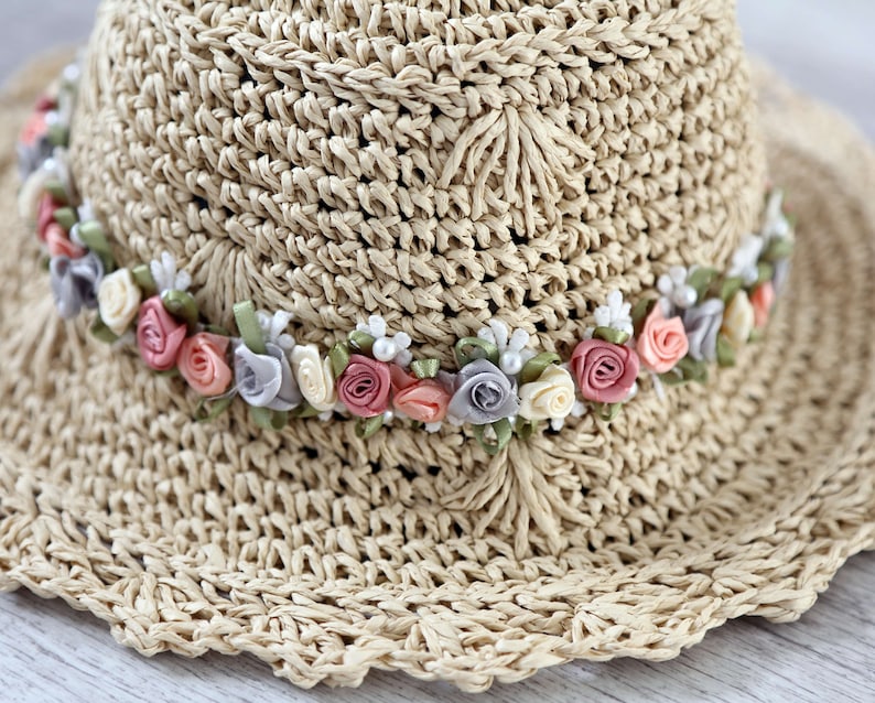 Puede incluir: Un sombrero de paja de ala ancha con una banda de flores de tela rosa, blanca y gris.
