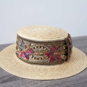 May include: A straw hat with a wide brim and a flat crown. The hat features a decorative band with floral embroidery in pink, blue, and gold sequins. The hat is a light beige color.