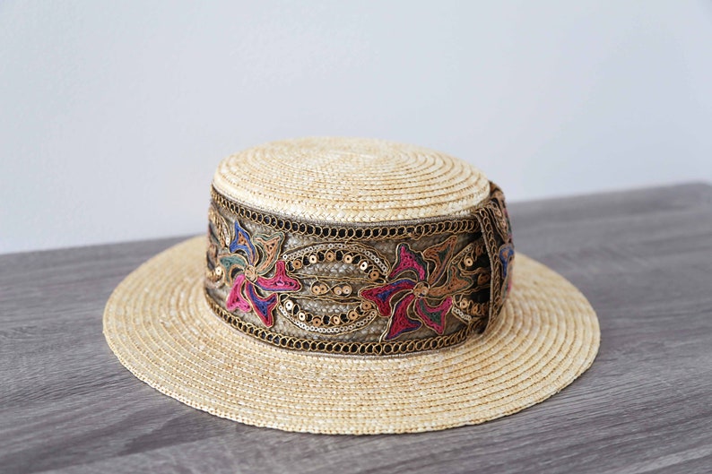 May include: A straw hat with a wide brim and a flat crown. The hat features a decorative band with floral embroidery in pink, blue, and gold sequins. The hat is a light beige color.