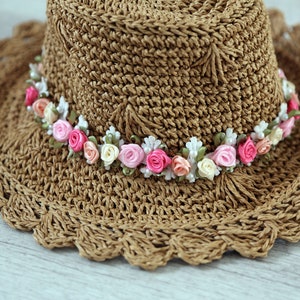 Puede incluir: Un sombrero de sol de crochet marr&oacute;n con un ala ancha y una banda de flores de tela rosa y blanca.