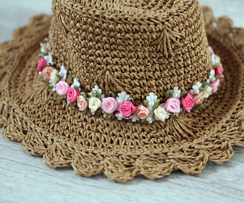 Puede incluir: Un sombrero de sol de crochet marr&oacute;n con un ala ancha y una banda de flores de tela rosa y blanca.