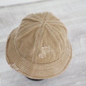 May include: A tan canvas bucket hat with white stitching and a distressed look. The hat has a small hole in the crown.