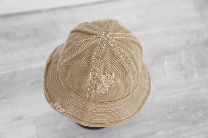 May include: A tan canvas bucket hat with white stitching and a distressed look. The hat has a small hole in the crown.
