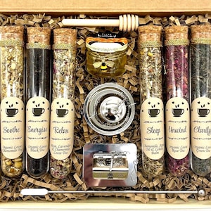 Op de afbeelding: Een cadeaupakket met glazen buizen gevuld met biologische losse thee, een klein potje honing, een thee-ei en een honingdipper. De buizen zijn gelabeld met theesoorten zoals "Soothe" en "Sleep". De set wordt gepresenteerd in een kartonnen doos.