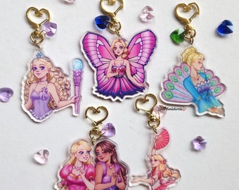May include: Five colorful acrylic charms featuring different characters from the Barbie Fairytopia movie series. Each charm has a gold heart-shaped ring and a clear acrylic background. The characters include Elina the Fairy, Nori the Fairy,  the Fairy Queen,  and two fairies holding hands.
