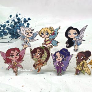 Op de afbeelding: Een verzameling van acht emaille pins met cartoonfeeën met kleurrijke vleugels en outfits. Elke pin heeft een unieke haarstijl en uitdrukking. De pins zijn geplaatst tegen een zachte, witte achtergrond met blauwe bloemaccenten.