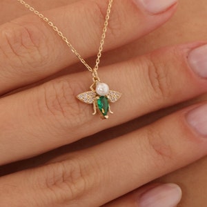 Puede incluir: Un collar de cadena dorada con un colgante de abeja verde y blanca. La abeja tiene un cuerpo de perla y alas verdes con piedras transparentes.
