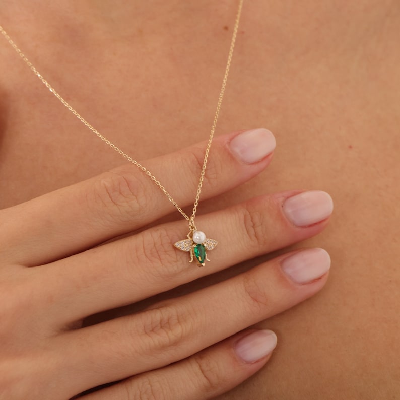 Puede incluir: Un collar de cadena dorada con un colgante de abeja verde y blanca. La abeja tiene un cuerpo de perla y alas verdes con piedras transparentes.