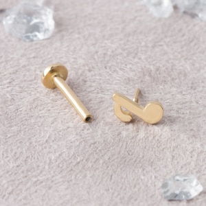 May include: Gold musical note stud earring with a post and a flat back. The earring is on a light brown surface.