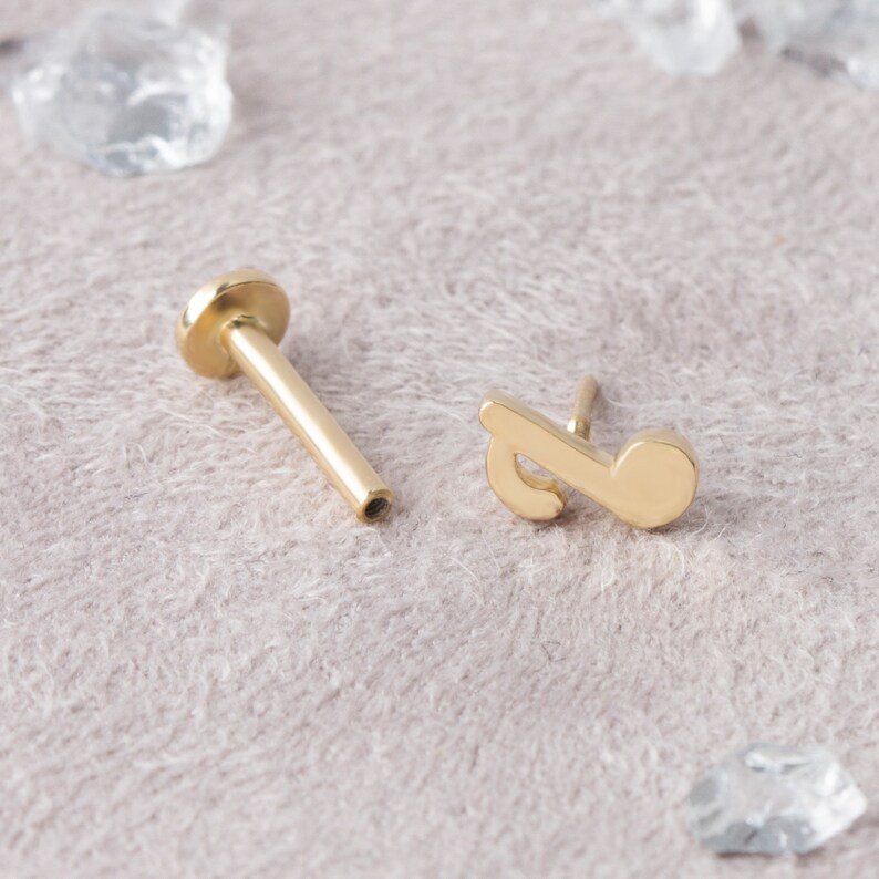 May include: Gold musical note stud earring with a post and a flat back. The earring is on a light brown surface.