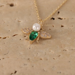 Puede incluir: Un collar de oro con un colgante de abeja. La abeja tiene una piedra preciosa verde y una perla blanca. La abeja est&aacute; rodeada de peque&ntilde;as piedras preciosas transparentes.