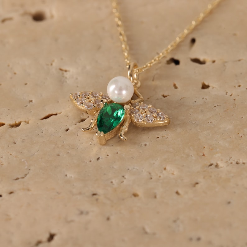 Puede incluir: Un collar de oro con un colgante de abeja. La abeja tiene una piedra preciosa verde y una perla blanca. La abeja est&aacute; rodeada de peque&ntilde;as piedras preciosas transparentes.