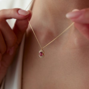 May include: A gold chain necklace with a pendant featuring a red oval gemstone surrounded by smaller clear gemstones.