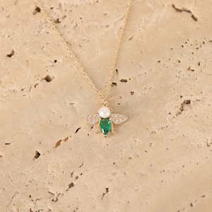 Puede incluir: Un collar de cadena dorada con un colgante de abeja. La abeja est&aacute; adornada con piedras preciosas transparentes y verdes y una perla blanca.