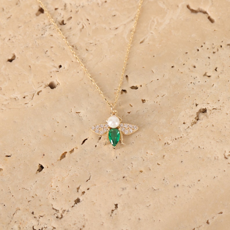 Puede incluir: Un collar de cadena dorada con un colgante de abeja. La abeja est&aacute; adornada con piedras preciosas transparentes y verdes y una perla blanca.