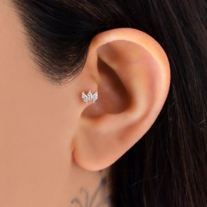 Pode incluir: Um close-up de uma orelha de uma pessoa com uma piercing de orelha de ouro e cristal transparente. O piercing tem a forma de uma flor com três pétalas.