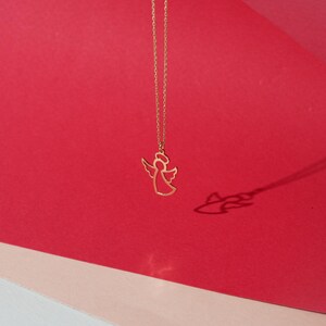 May include: A gold chain necklace with a delicate angel pendant. The angel is outlined in gold and has a halo. The necklace is photographed against a red and pink background.