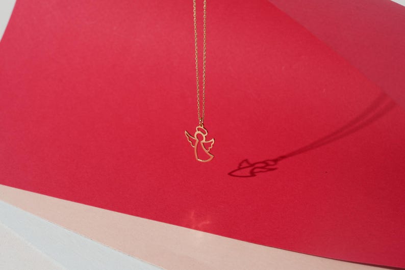 May include: A gold chain necklace with a delicate angel pendant. The angel is outlined in gold and has a halo. The necklace is photographed against a red and pink background.