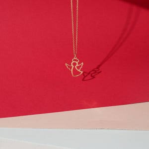 May include: A gold angel pendant necklace on a red and pink background.