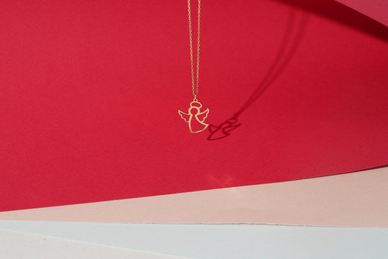 May include: A gold angel pendant necklace on a red and pink background.